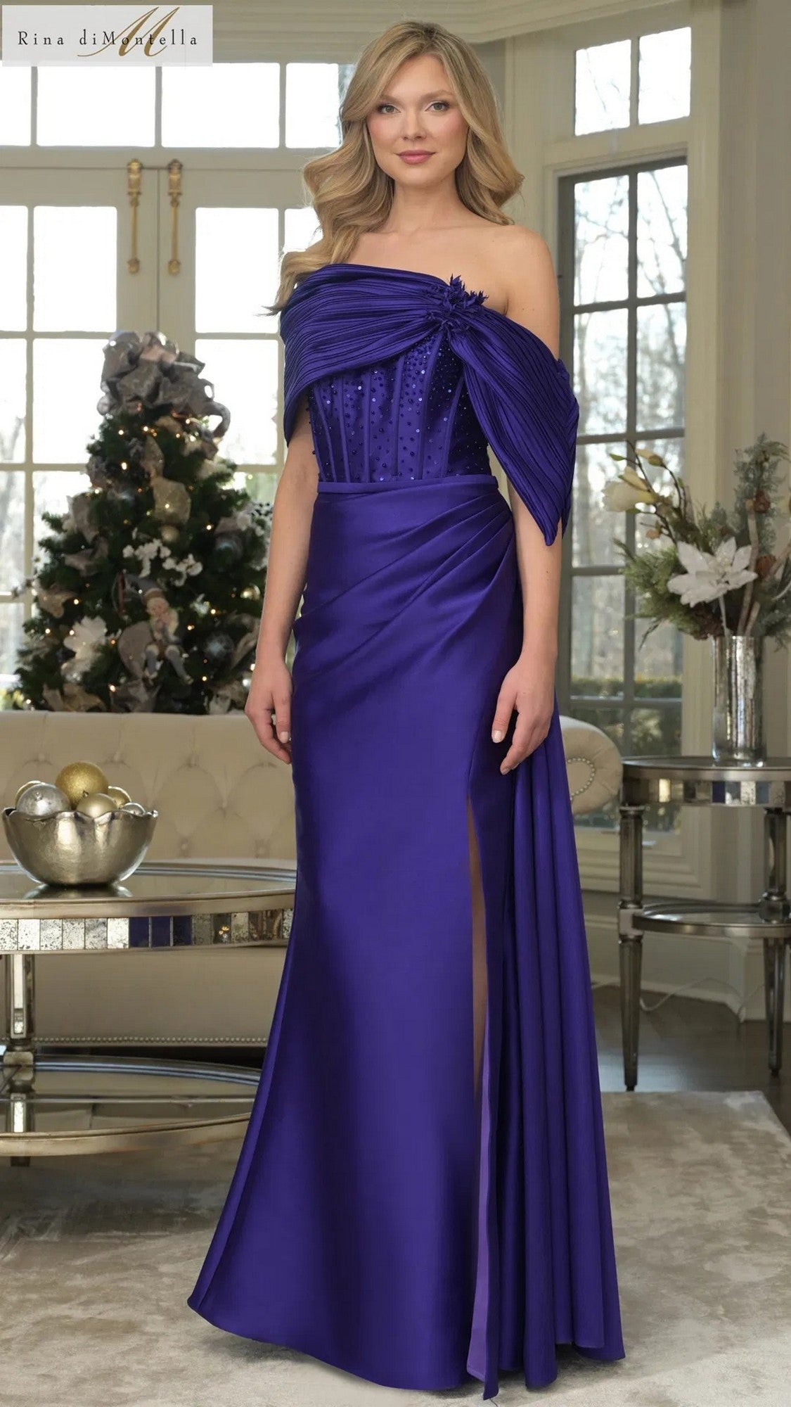 A woman indoors wears the amethyst Long Formal Dress: Rina Di Montella RD3309, a floor-length off-shoulder gown with sheath skirt and side slit. A decorated Christmas tree and sofa are in the background.